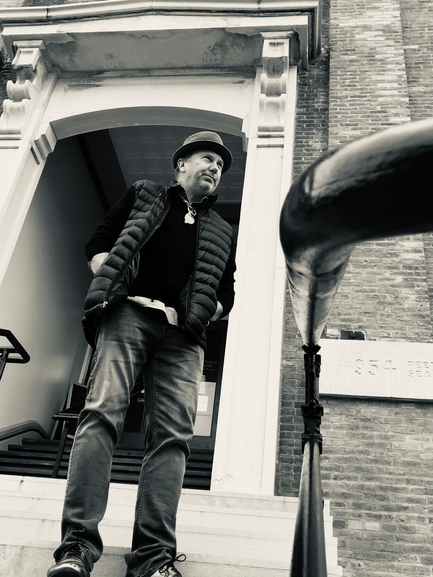 John Kirkpatrick in a brimmed hat and vest, standing at the top of an exterior staircase in front of an arched entrance